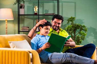 Hintli Asyalı oğlu babasıyla birlikte modern evdeki kanepede kitap okurken okul çocuğu özenle ders çalışıyor ve babası da rahat oturma odasında aile eğitimi anlarında öğrenmeyi destekliyor.
