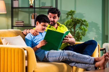 Hintli Asyalı oğlu babasıyla birlikte modern evdeki kanepede kitap okurken okul çocuğu özenle ders çalışıyor ve babası da rahat oturma odasında aile eğitimi anlarında öğrenmeyi destekliyor.