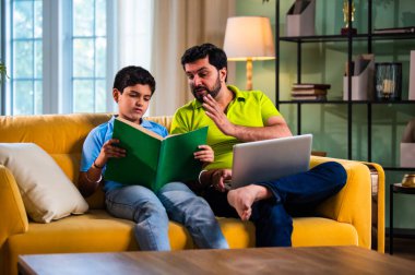 Hintli Asyalı oğlu kitap okuyor babası modern evde dizüstü bilgisayarda çalışırken okul çocuğu kanepede çalışıyor ve baba oturma odasında sakin aile öğrenme süresince şüpheleri cevaplıyor.