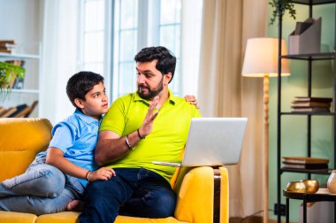 Hintli genç Asyalı baba oğul, modern evde dizüstü bilgisayar kullanırken kanepeye oturmuş internette alışveriş yapıyor ve şık oturma odasında dijital bağ kurmanın keyfini çıkarıyor.