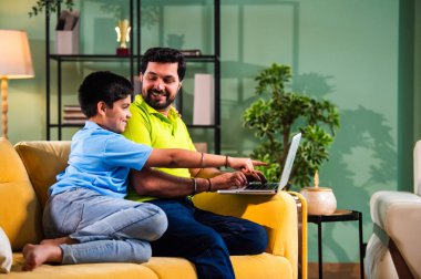 Hintli genç Asyalı baba oğul, modern evde dizüstü bilgisayar kullanırken kanepeye oturmuş internette alışveriş yapıyor ve şık oturma odasında dijital bağ kurmanın keyfini çıkarıyor.