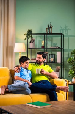 Hintli Asyalı baba oğul, modern evdeki koltukta oturmuş sohbet ederken neşeli anları paylaşıyor, sıcak aile bağları ve oturma odasındaki kaliteli zaman sırasında birlikte gülüyor ve gülümsüyor.