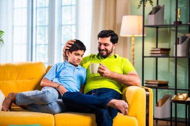 Hintli Asyalı baba oğul, modern evdeki koltukta oturmuş sohbet ederken neşeli anları paylaşıyor, sıcak aile bağları ve oturma odasındaki kaliteli zaman sırasında birlikte gülüyor ve gülümsüyor.