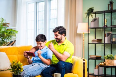 Hintli Asyalı genç baba oğlu modern evdeki koltukta oturup eğlenceli dijital aktivitelerin tadını çıkarırken oturma odasında rahat ve kaliteli bir aile zamanı geçirirken gülümsüyor ve bağ kuruyor.