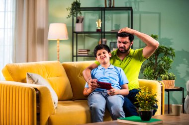 Hintli Asyalı genç baba oğlu modern evdeki koltukta oturup eğlenceli dijital aktivitelerin tadını çıkarırken oturma odasında rahat ve kaliteli bir aile zamanı geçirirken gülümsüyor ve bağ kuruyor.