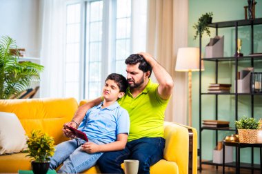 Hintli Asyalı genç baba oğlu modern evdeki koltukta oturup eğlenceli dijital aktivitelerin tadını çıkarırken oturma odasında rahat ve kaliteli bir aile zamanı geçirirken gülümsüyor ve bağ kuruyor.