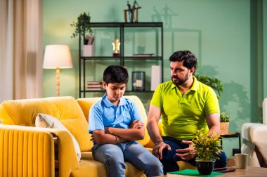 Hintli genç baba, modern evdeki koltukta otururken oğlunu azarlıyor. Üzgün çocuk sessizce arkasını dönüyor. Oturma odasında duygusal ebeveynlik anı ve aile gerginliği yaşıyor.