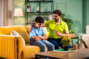 Hintli genç baba, modern evdeki koltukta otururken oğlunu azarlıyor. Üzgün çocuk sessizce arkasını dönüyor. Oturma odasında duygusal ebeveynlik anı ve aile gerginliği yaşıyor.