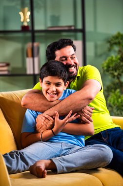 Hintli mutlu genç baba, modern evdeki koltukta oturup sevgi dolu aile bağlarının tadını çıkarırken sıcak oturma odasında sevgi, yakınlık ve rahatlık göstererek oğluna sarılıyor.