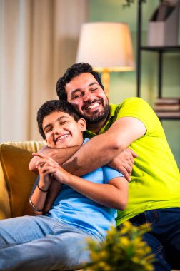 Hintli mutlu genç baba, modern evdeki koltukta oturup sevgi dolu aile bağlarının tadını çıkarırken sıcak oturma odasında sevgi, yakınlık ve rahatlık göstererek oğluna sarılıyor.