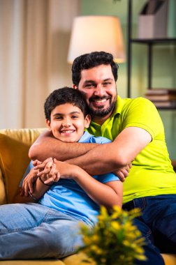 Hintli mutlu genç baba, modern evdeki koltukta oturup sevgi dolu aile bağlarının tadını çıkarırken sıcak oturma odasında sevgi, yakınlık ve rahatlık göstererek oğluna sarılıyor.