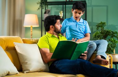 Hintli baba ve oğlu modern evdeki kanepede kitap okurken, baba okul bölümünü anlatıyor ve küçük çocuğa sıcak oturma odasında oturup aile eğitimi anlarını anlatıyor.