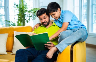 Hintli baba ve oğlu modern evdeki kanepede kitap okurken, baba okul bölümünü anlatıyor ve küçük çocuğa sıcak oturma odasında oturup aile eğitimi anlarını anlatıyor.