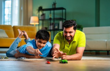 Hintli Asyalı genç baba oğul oyuncak araba yarışı oynarken spor müsabakalarının tadını çıkarıyor, modern oturma odasında eğlenirken ve kaynaşırken gülüyor ve kaynaşıyor.