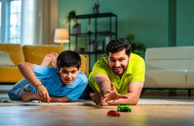 Hintli Asyalı genç baba oğul oyuncak araba yarışı oynarken spor müsabakalarının tadını çıkarıyor, modern oturma odasında eğlenirken ve kaynaşırken gülüyor ve kaynaşıyor.