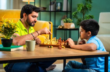 Hintli genç baba oğul evde satranç oynuyor arkadaşça rekabet ve spor rekabetinin tadını çıkarıyor, birlikte strateji öğrenirken modern oturma odasında kaliteli aile zamanı geçiriyor.