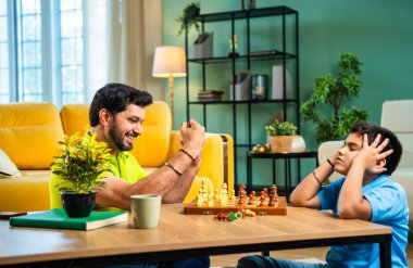 Hintli genç baba oğul evde satranç oynuyor arkadaşça rekabet ve spor rekabetinin tadını çıkarıyor, birlikte strateji öğrenirken modern oturma odasında kaliteli aile zamanı geçiriyor.