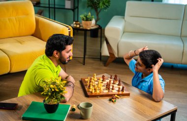 Hintli genç baba oğul evde satranç oynuyor arkadaşça rekabet ve spor rekabetinin tadını çıkarıyor, birlikte strateji öğrenirken modern oturma odasında kaliteli aile zamanı geçiriyor.