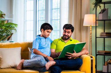 Hintli baba ve oğlu modern evdeki kanepede kitap okurken, baba okul bölümünü anlatıyor ve küçük çocuğa sıcak oturma odasında oturup aile eğitimi anlarını anlatıyor.