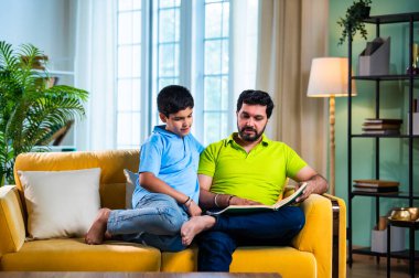 Hintli baba ve oğlu modern evdeki kanepede kitap okurken, baba okul bölümünü anlatıyor ve küçük çocuğa sıcak oturma odasında oturup aile eğitimi anlarını anlatıyor.