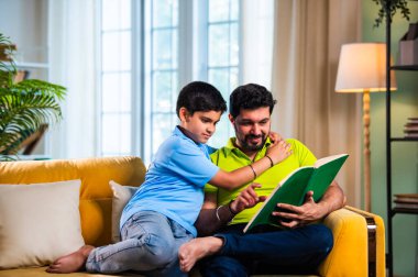 Hintli baba ve oğlu modern evdeki kanepede kitap okurken, baba okul bölümünü anlatıyor ve küçük çocuğa sıcak oturma odasında oturup aile eğitimi anlarını anlatıyor.