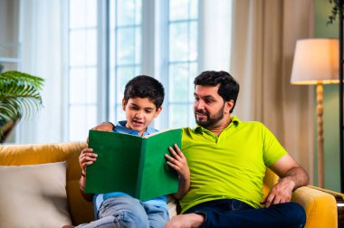 Hintli Asyalı oğlu babasıyla birlikte modern evdeki kanepede kitap okurken okul çocuğu özenle ders çalışıyor ve babası da rahat oturma odasında aile eğitimi anlarında öğrenmeyi destekliyor.