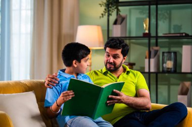 Hintli Asyalı oğlu babasıyla birlikte modern evdeki kanepede kitap okurken okul çocuğu özenle ders çalışıyor ve babası da rahat oturma odasında aile eğitimi anlarında öğrenmeyi destekliyor.
