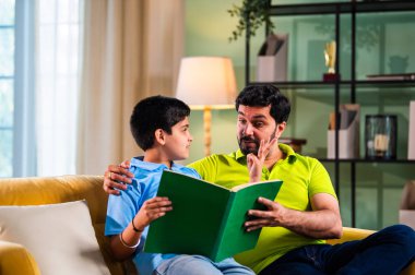 Hintli Asyalı oğlu babasıyla birlikte modern evdeki kanepede kitap okurken okul çocuğu özenle ders çalışıyor ve babası da rahat oturma odasında aile eğitimi anlarında öğrenmeyi destekliyor.