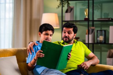 Hintli Asyalı oğlu babasıyla birlikte modern evdeki kanepede kitap okurken okul çocuğu özenle ders çalışıyor ve babası da rahat oturma odasında aile eğitimi anlarında öğrenmeyi destekliyor.