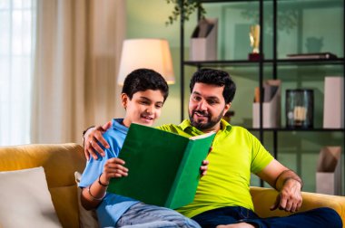 Hintli Asyalı oğlu babasıyla birlikte modern evdeki kanepede kitap okurken okul çocuğu özenle ders çalışıyor ve babası da rahat oturma odasında aile eğitimi anlarında öğrenmeyi destekliyor.