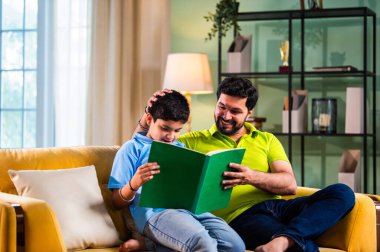 Hintli Asyalı oğlu babasıyla birlikte modern evdeki kanepede kitap okurken okul çocuğu özenle ders çalışıyor ve babası da rahat oturma odasında aile eğitimi anlarında öğrenmeyi destekliyor.