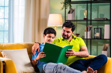 Hintli Asyalı oğlu babasıyla birlikte modern evdeki kanepede kitap okurken okul çocuğu özenle ders çalışıyor ve babası da rahat oturma odasında aile eğitimi anlarında öğrenmeyi destekliyor.