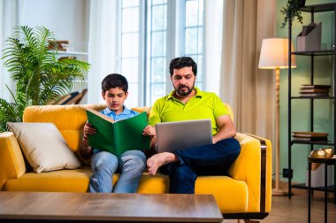 Hintli Asyalı oğlu kitap okuyor babası modern evde dizüstü bilgisayarda çalışırken okul çocuğu kanepede çalışıyor ve baba oturma odasında sakin aile öğrenme süresince şüpheleri cevaplıyor.