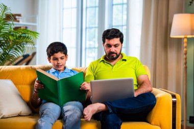 Hintli Asyalı oğlu kitap okuyor babası modern evde dizüstü bilgisayarda çalışırken okul çocuğu kanepede çalışıyor ve baba oturma odasında sakin aile öğrenme süresince şüpheleri cevaplıyor.