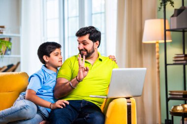 Hintli genç Asyalı baba oğul, modern evde dizüstü bilgisayar kullanırken kanepeye oturmuş internette alışveriş yapıyor ve şık oturma odasında dijital bağ kurmanın keyfini çıkarıyor.