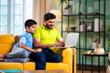 Hintli genç Asyalı baba oğul, modern evde dizüstü bilgisayar kullanırken kanepeye oturmuş internette alışveriş yapıyor ve şık oturma odasında dijital bağ kurmanın keyfini çıkarıyor.