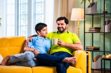 Hintli Asyalı baba oğul, modern evdeki koltukta oturmuş sohbet ederken neşeli anları paylaşıyor, sıcak aile bağları ve oturma odasındaki kaliteli zaman sırasında birlikte gülüyor ve gülümsüyor.