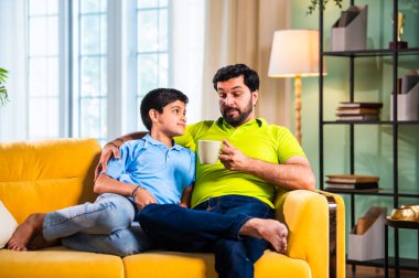 Hintli Asyalı baba oğul, modern evdeki koltukta oturmuş sohbet ederken neşeli anları paylaşıyor, sıcak aile bağları ve oturma odasındaki kaliteli zaman sırasında birlikte gülüyor ve gülümsüyor.