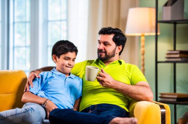 Hintli Asyalı baba oğul, modern evdeki koltukta oturmuş sohbet ederken neşeli anları paylaşıyor, sıcak aile bağları ve oturma odasındaki kaliteli zaman sırasında birlikte gülüyor ve gülümsüyor.