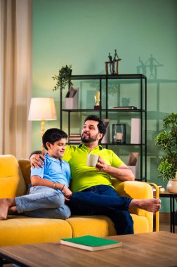 Hintli Asyalı baba oğul, modern evdeki koltukta oturmuş sohbet ederken neşeli anları paylaşıyor, sıcak aile bağları ve oturma odasındaki kaliteli zaman sırasında birlikte gülüyor ve gülümsüyor.