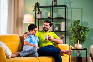 Hintli Asyalı baba oğul, modern evdeki koltukta oturmuş sohbet ederken neşeli anları paylaşıyor, sıcak aile bağları ve oturma odasındaki kaliteli zaman sırasında birlikte gülüyor ve gülümsüyor.