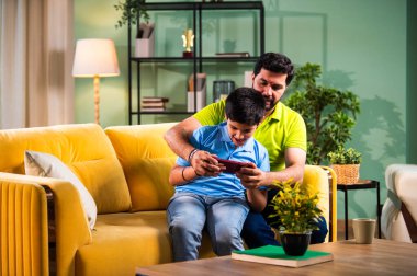 Hintli Asyalı genç baba oğlu modern evdeki koltukta oturup eğlenceli dijital aktivitelerin tadını çıkarırken oturma odasında rahat ve kaliteli bir aile zamanı geçirirken gülümsüyor ve bağ kuruyor.