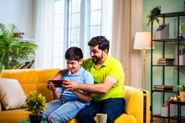 Hintli Asyalı genç baba oğlu modern evdeki koltukta oturup eğlenceli dijital aktivitelerin tadını çıkarırken oturma odasında rahat ve kaliteli bir aile zamanı geçirirken gülümsüyor ve bağ kuruyor.
