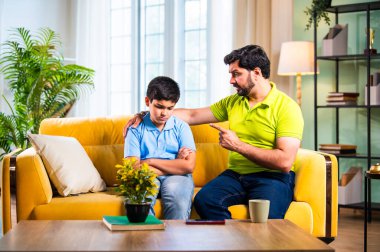 Hintli genç baba, modern evdeki koltukta otururken oğlunu azarlıyor. Üzgün çocuk sessizce arkasını dönüyor. Oturma odasında duygusal ebeveynlik anı ve aile gerginliği yaşıyor.