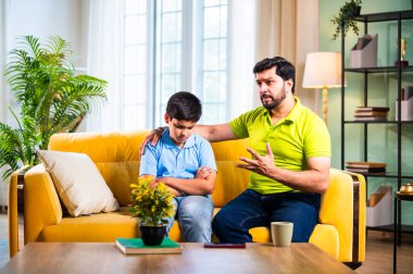 Hintli genç baba, modern evdeki koltukta otururken oğlunu azarlıyor. Üzgün çocuk sessizce arkasını dönüyor. Oturma odasında duygusal ebeveynlik anı ve aile gerginliği yaşıyor.