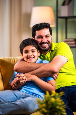 Hintli mutlu genç baba, modern evdeki koltukta oturup sevgi dolu aile bağlarının tadını çıkarırken sıcak oturma odasında sevgi, yakınlık ve rahatlık göstererek oğluna sarılıyor.
