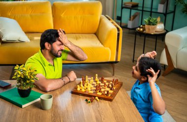 Hintli genç baba oğul evde satranç oynuyor arkadaşça rekabet ve spor rekabetinin tadını çıkarıyor, birlikte strateji öğrenirken modern oturma odasında kaliteli aile zamanı geçiriyor.