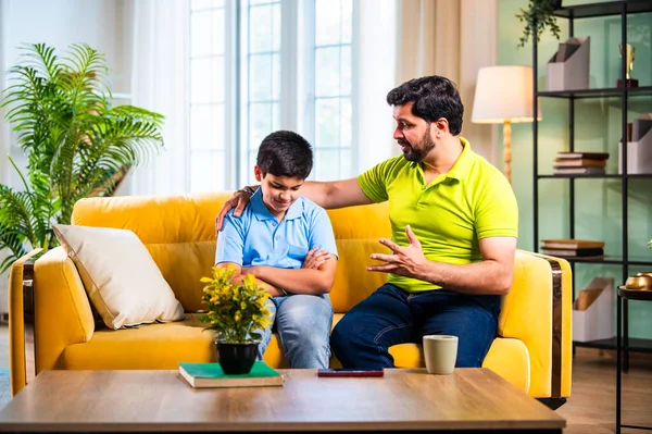 Hintli genç baba, modern evdeki koltukta otururken oğlunu azarlıyor. Üzgün çocuk sessizce arkasını dönüyor. Oturma odasında duygusal ebeveynlik anı ve aile gerginliği yaşıyor.