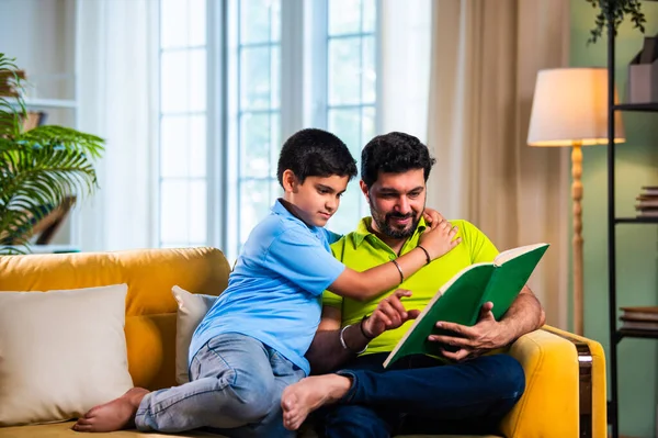 Hintli baba ve oğlu modern evdeki kanepede kitap okurken, baba okul bölümünü anlatıyor ve küçük çocuğa sıcak oturma odasında oturup aile eğitimi anlarını anlatıyor.