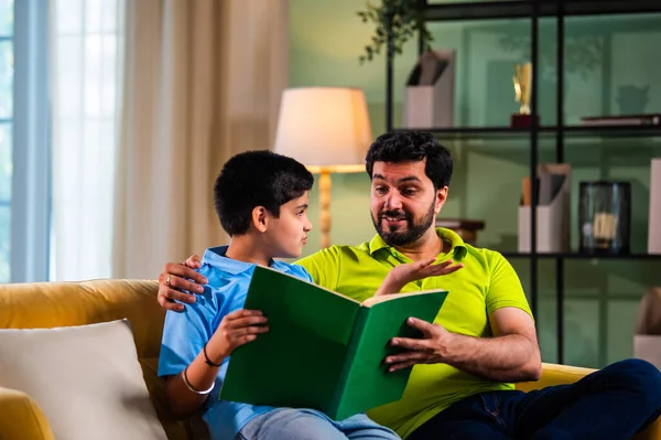 Hintli Asyalı oğlu babasıyla birlikte modern evdeki kanepede kitap okurken okul çocuğu özenle ders çalışıyor ve babası da rahat oturma odasında aile eğitimi anlarında öğrenmeyi destekliyor.